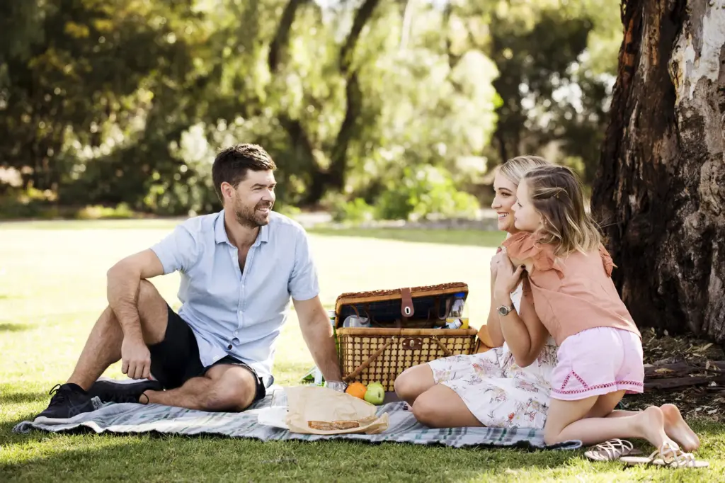 A man, woman, and young girl enjoy a picnic on a blanket in a sunny park. They are sitting near a picnic basket with food, surrounded by trees and green grass, smiling and talking together.