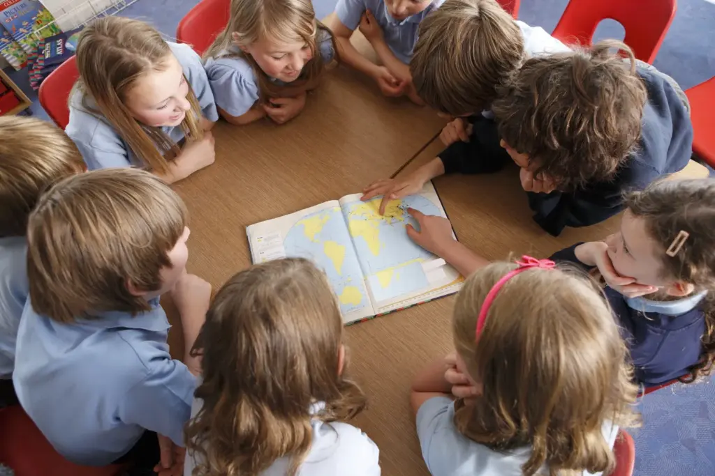 A group of children sit around a table looking at an open world map, with one child pointing at a location, suggesting a group learning or geography activity.