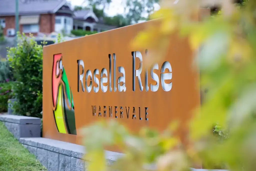A large rust-colored metal sign reads "Rosella Rise Warnervale," with a colorful bird illustration on the left. Greenery and blurred leaves frame the sign; houses are visible in the background.